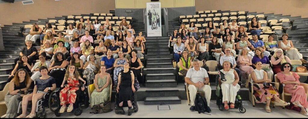Un grupo de mujeres y hombres sentado en un salón de actos. En primera línea, al menos hay dos mujeres en silla de ruedas