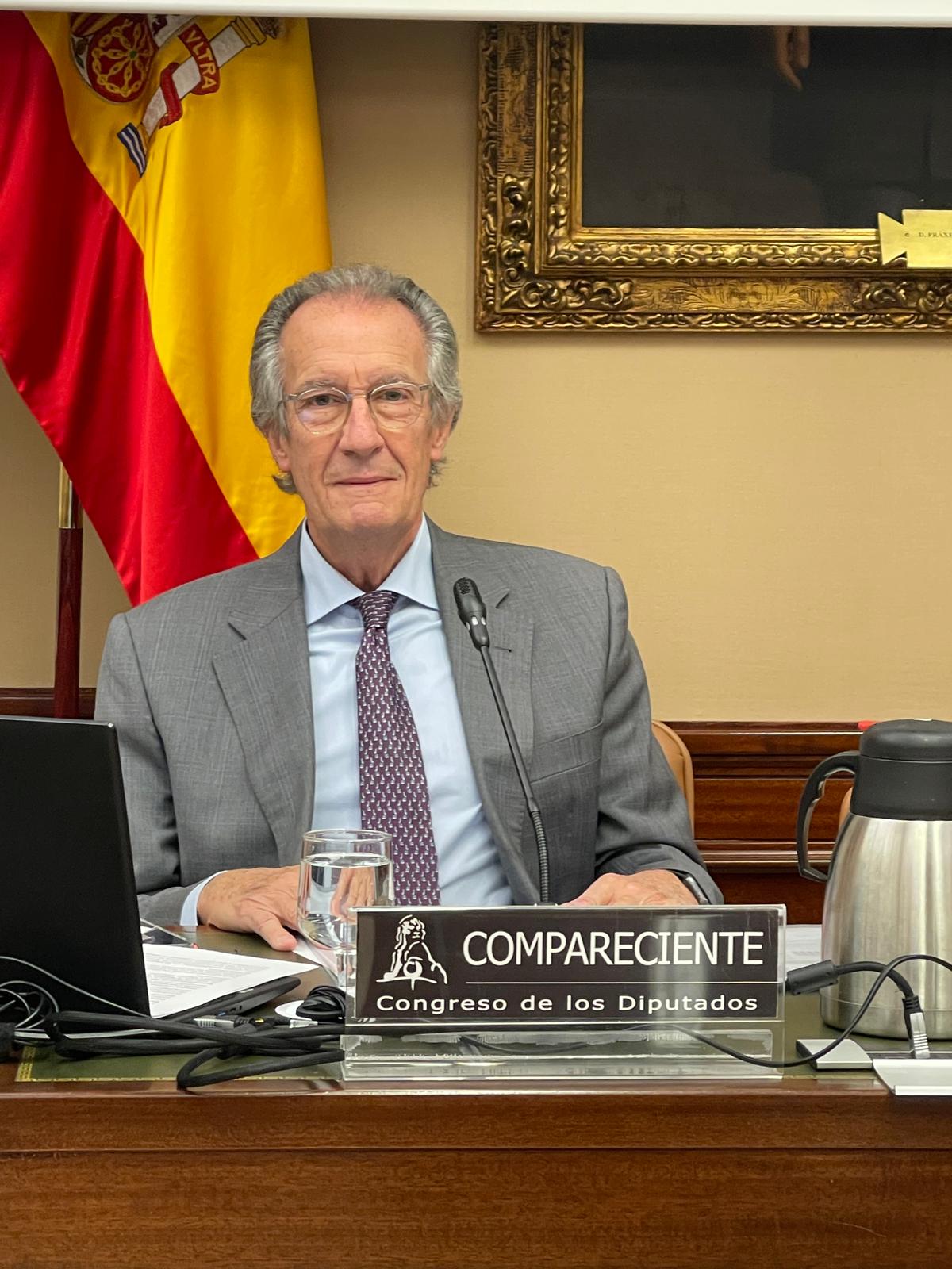 Un hombre mayor, vestido de traje y con gafas, sentado en una mesa institucional tras el cartel de compareciente.